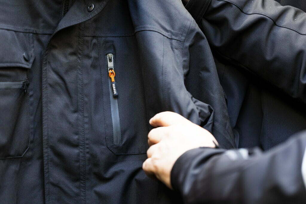 Hånd som rører ved en svart arbeidsjakke med oransje glidelåssnor.